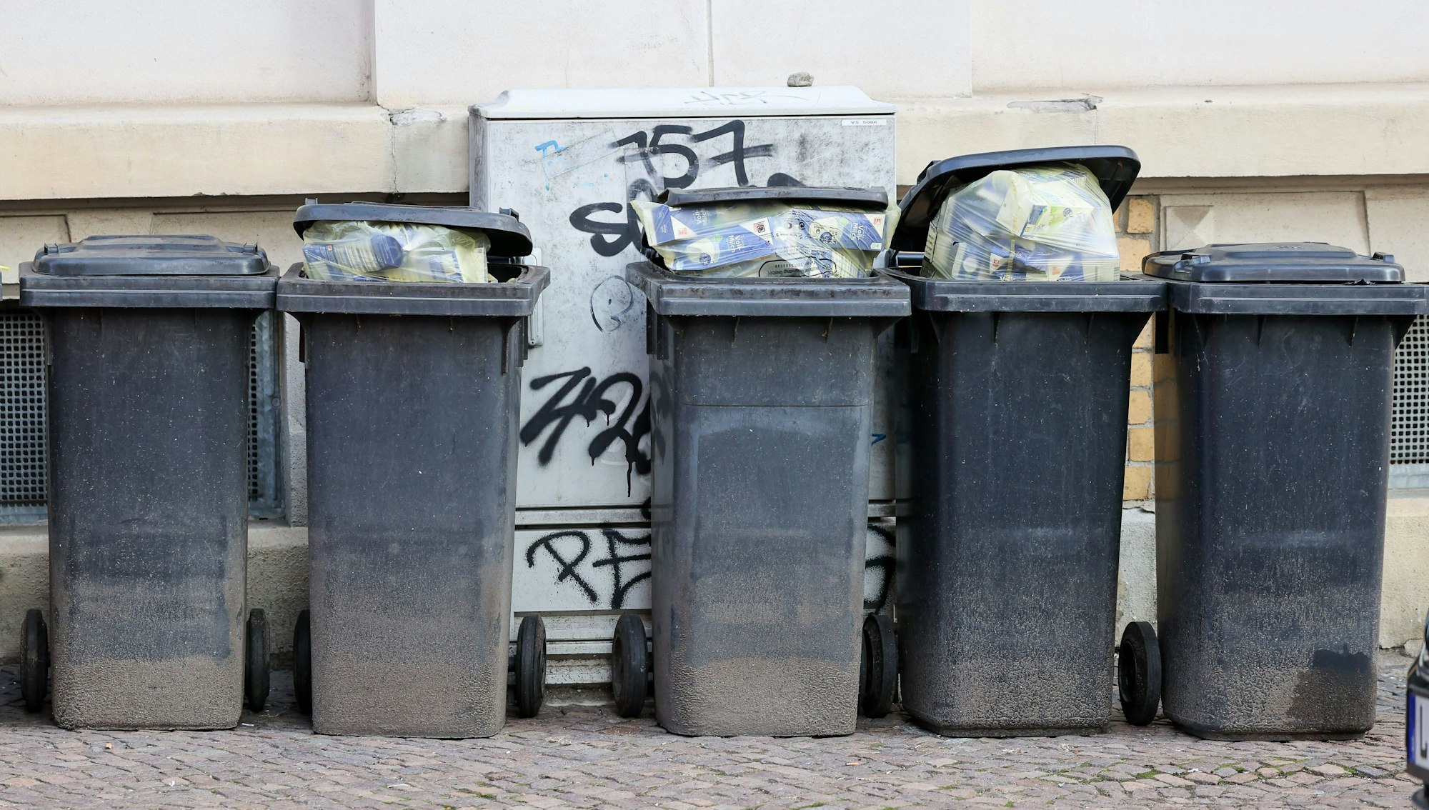 Mehrere volle Mülltonnen für Restmüll stehen an einer Hauswand.