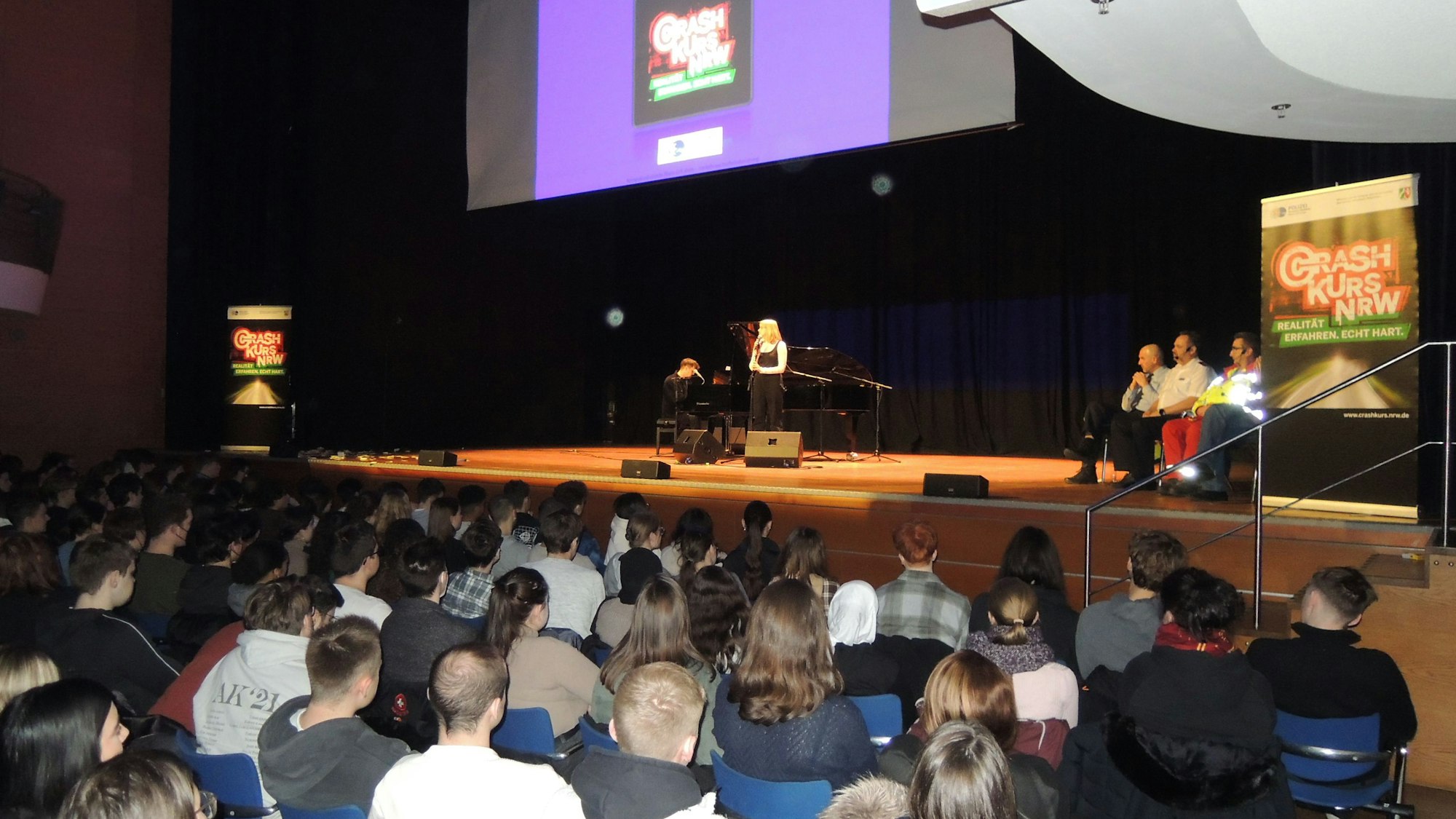 Jugendliche sitzen in einem Forum vor einer Bühne, auf der Aufsteller mit „Crash Kurs NRW“ stehen und einige Leute sitzen.