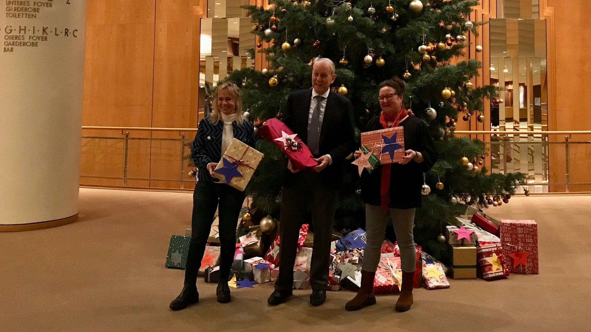 Gaby Schauenburg, Louwrens Langevoort und Gabi Schaume mit Geschenken in der Hand vor einem geschmückten Weihnachtsbaum mit Geschenken