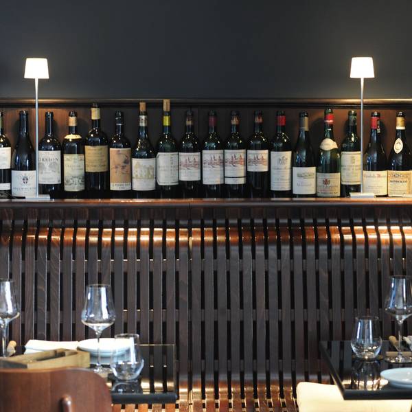 Beleuchtetes Regal mit Weinflaschen, davor gedeckte Tische eines Restaurants.