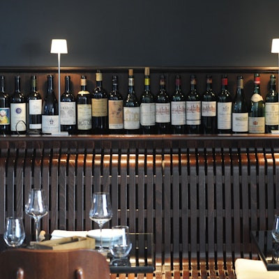 Beleuchtetes Regal mit Weinflaschen, davor gedeckte Tische eines Restaurants.