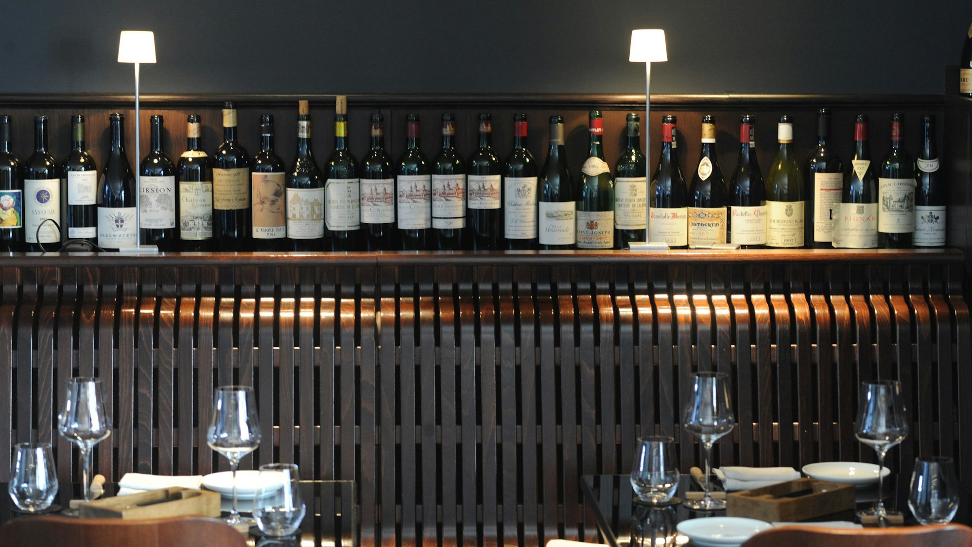 Beleuchtetes Regal mit Weinflaschen, davor gedeckte Tische eines Restaurants.
