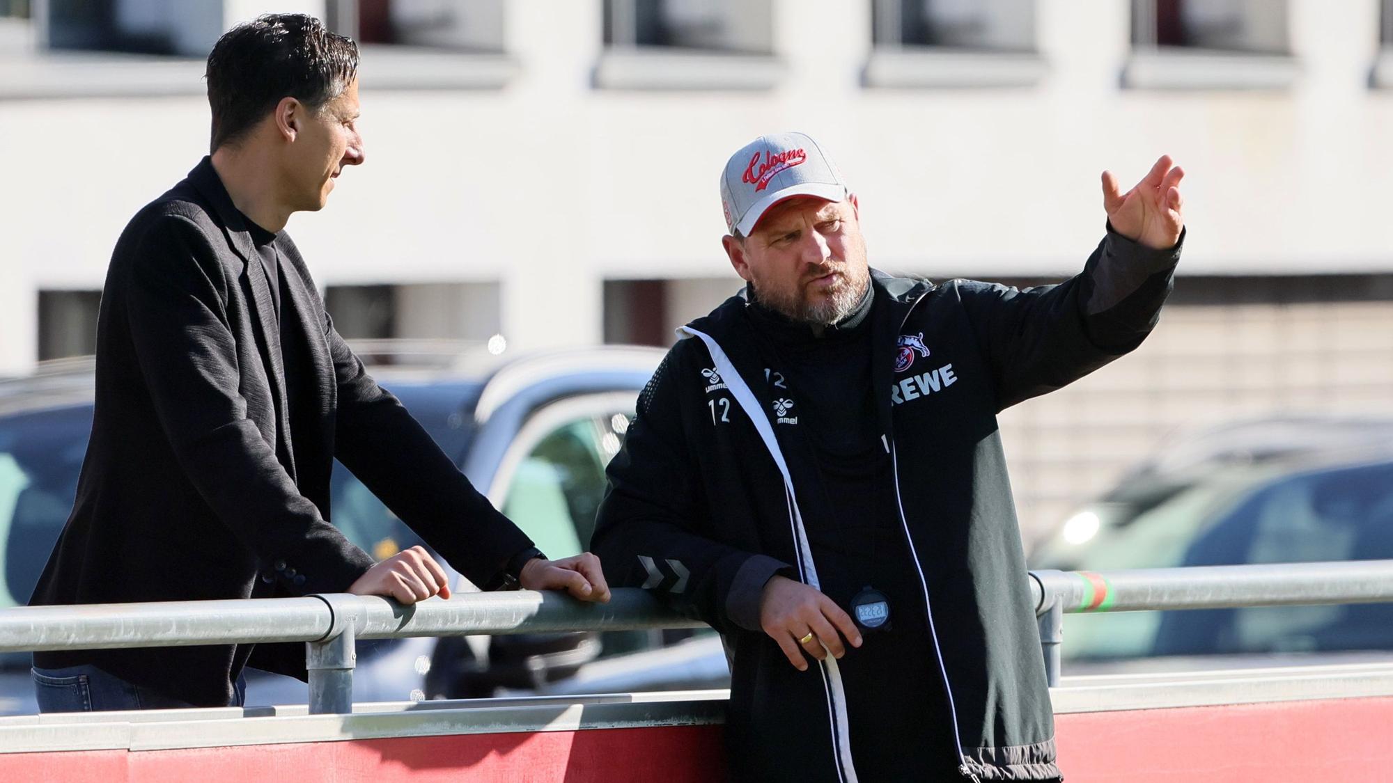 FC-Geschäftsführer Christian Keller und Trainer Steffen Baumgart unterhalten sich am Rande des Trainings des 1. FC Köln am Geißbockheim. Baumgart lehnt an der Bande und gestikuliert, Keller steht dahinter.