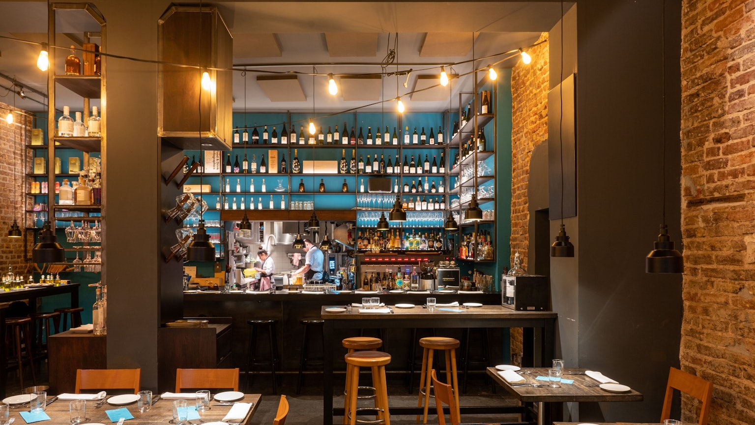 Innenraum eines Restaurants mit Holzstühlen und -tischen, im Hintergrund eine Bar