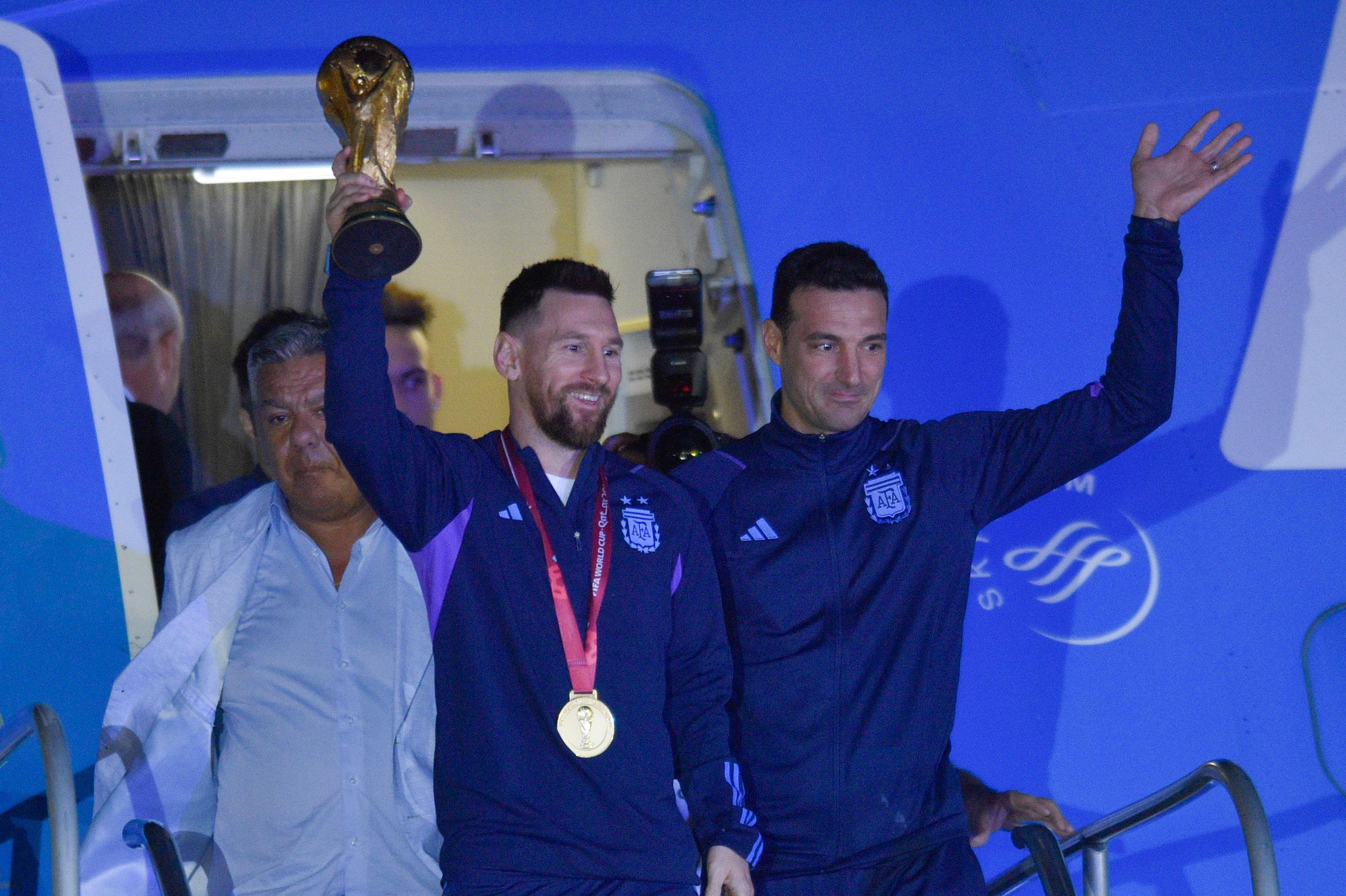 Lionel Messi steht auf einer Flugzeugtreppe vor einem Flugzeug und reckt den Weltmeisterpokal mit einer Hand in die Höhe.