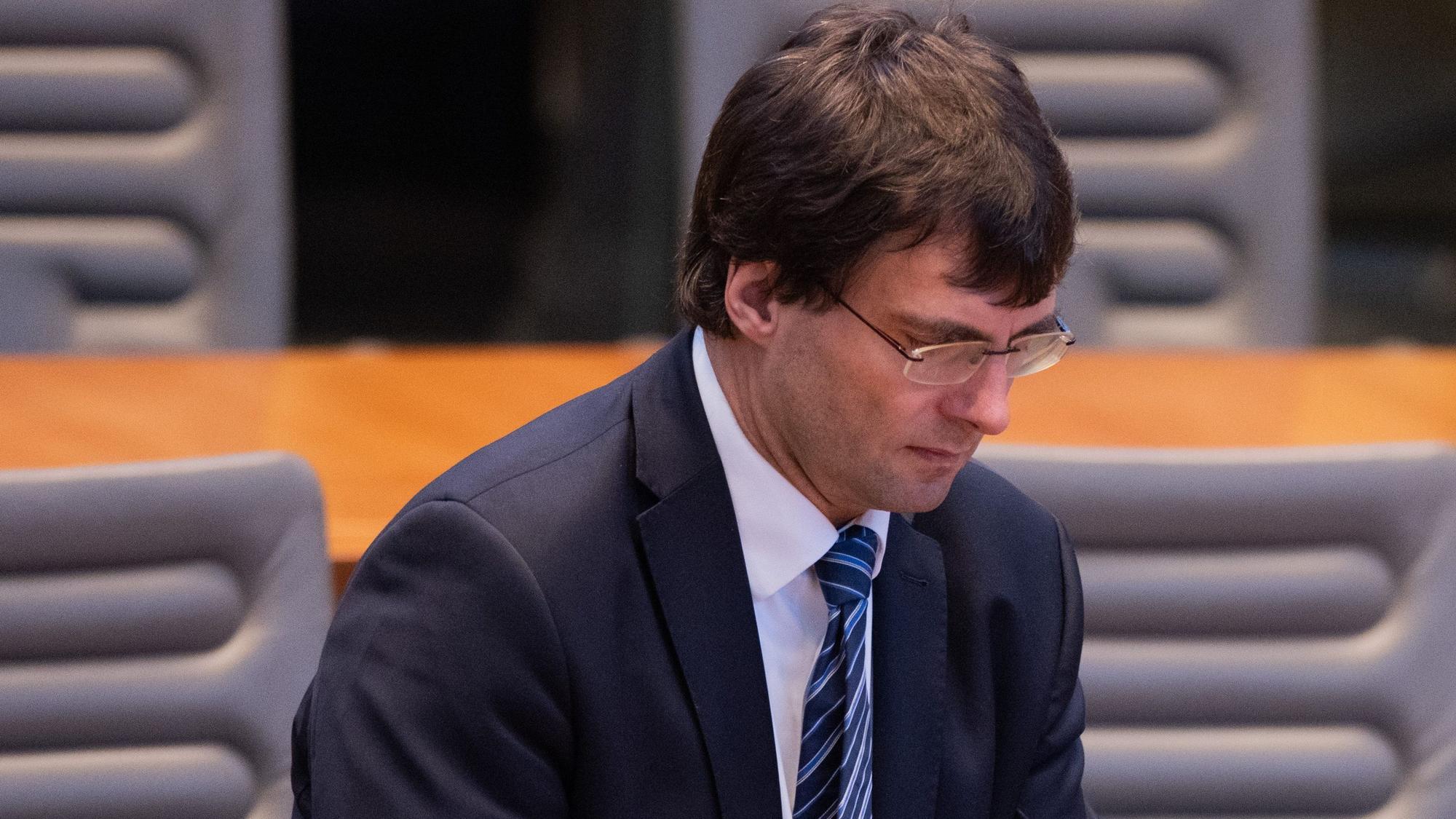 Minister Optendrenk ist im Plenum zu sehen, wie er seinen Stuhl im Stehen zurechtrückt. Er blickt versonnen nach unten.