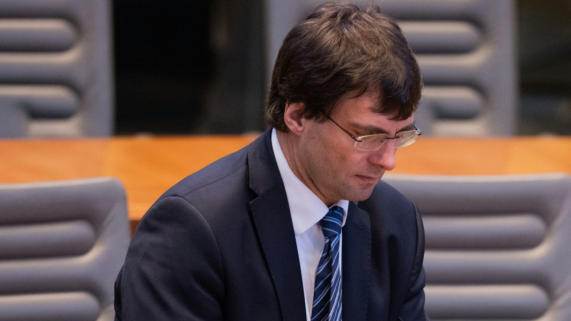 Minister Optendrenk ist im Plenum zu sehen, wie er seinen Stuhl im Stehen zurechtrückt. Er blickt versonnen nach unten.