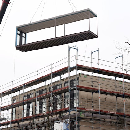 Ein Kran liefert ein Modul für eine Schule an. Die Schule ist mit einem rot-silbernen Gerüst eingerüstet.