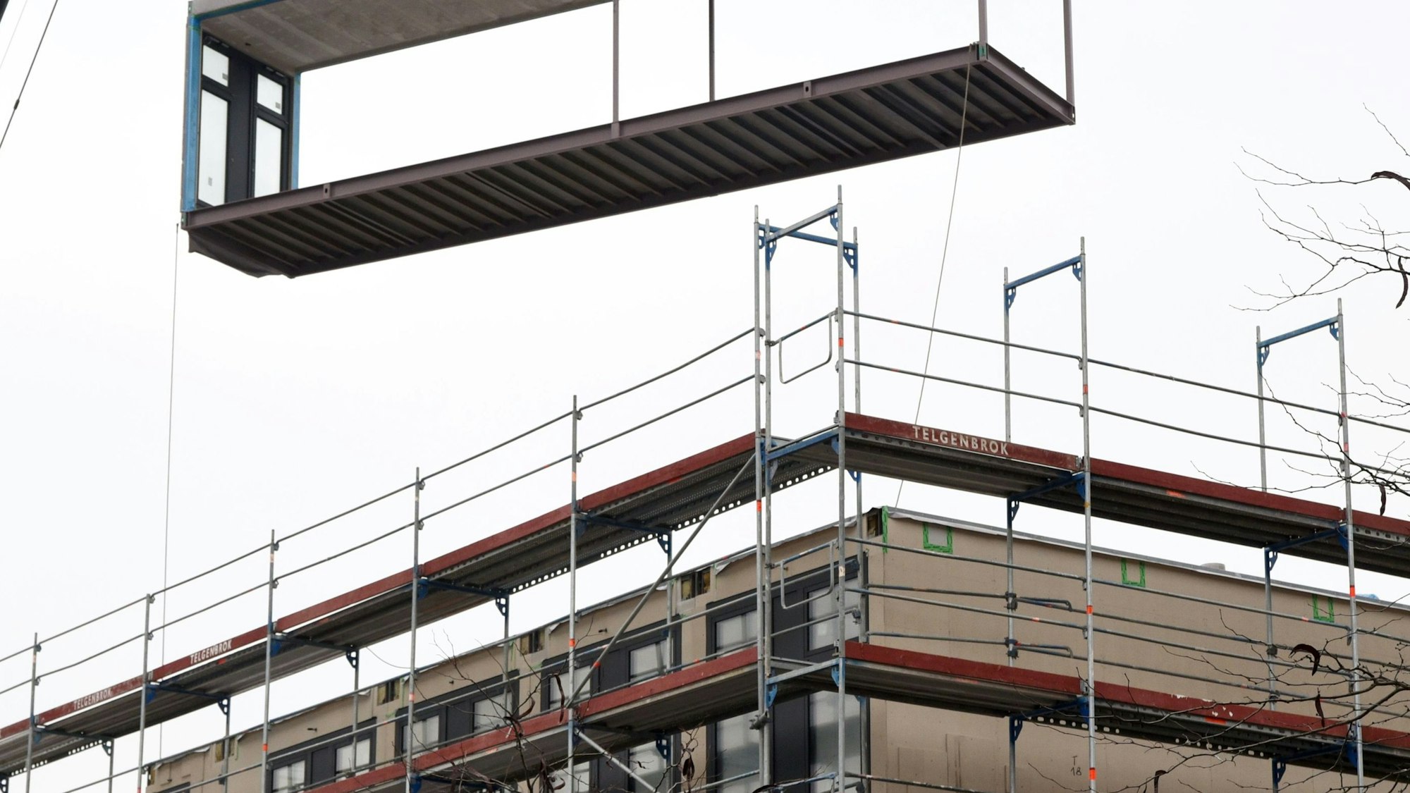 Ein Kran liefert ein Modul für eine Schule an. Die Schule ist mit einem rot-silbernen Gerüst eingerüstet.