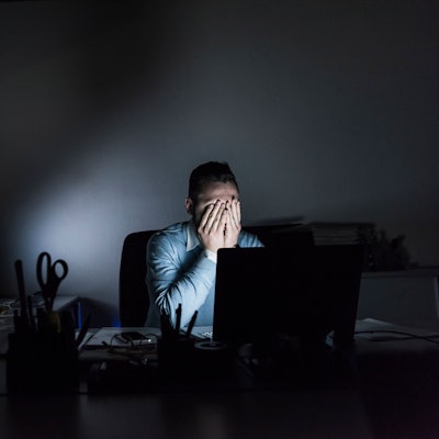 Ein Mann sitzt in einem dunklen Büro am Computer und hält sich die Hände vors Gesicht.