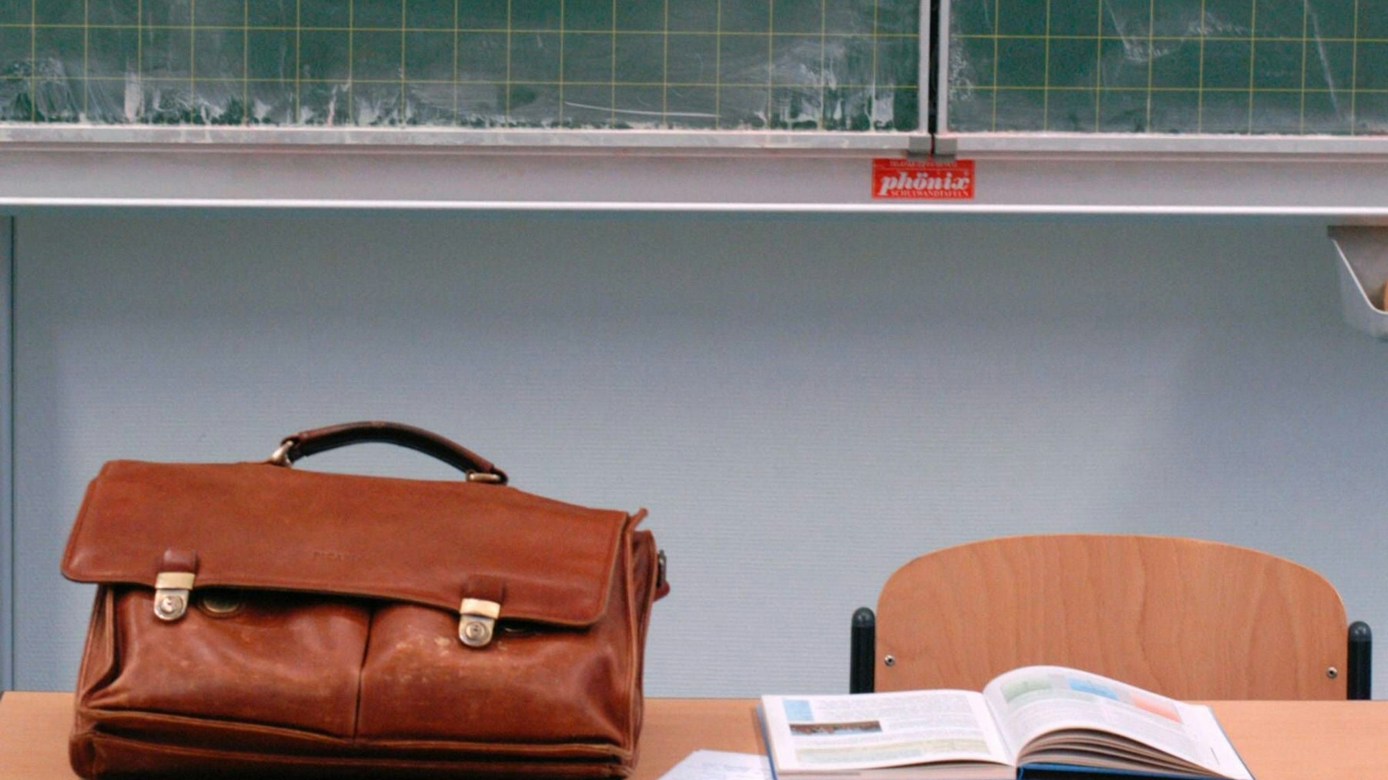 Auf einem Pult liegt eine braune Lehrertasche. Im Hintergrund ist eine leere Tafel zu sehen.