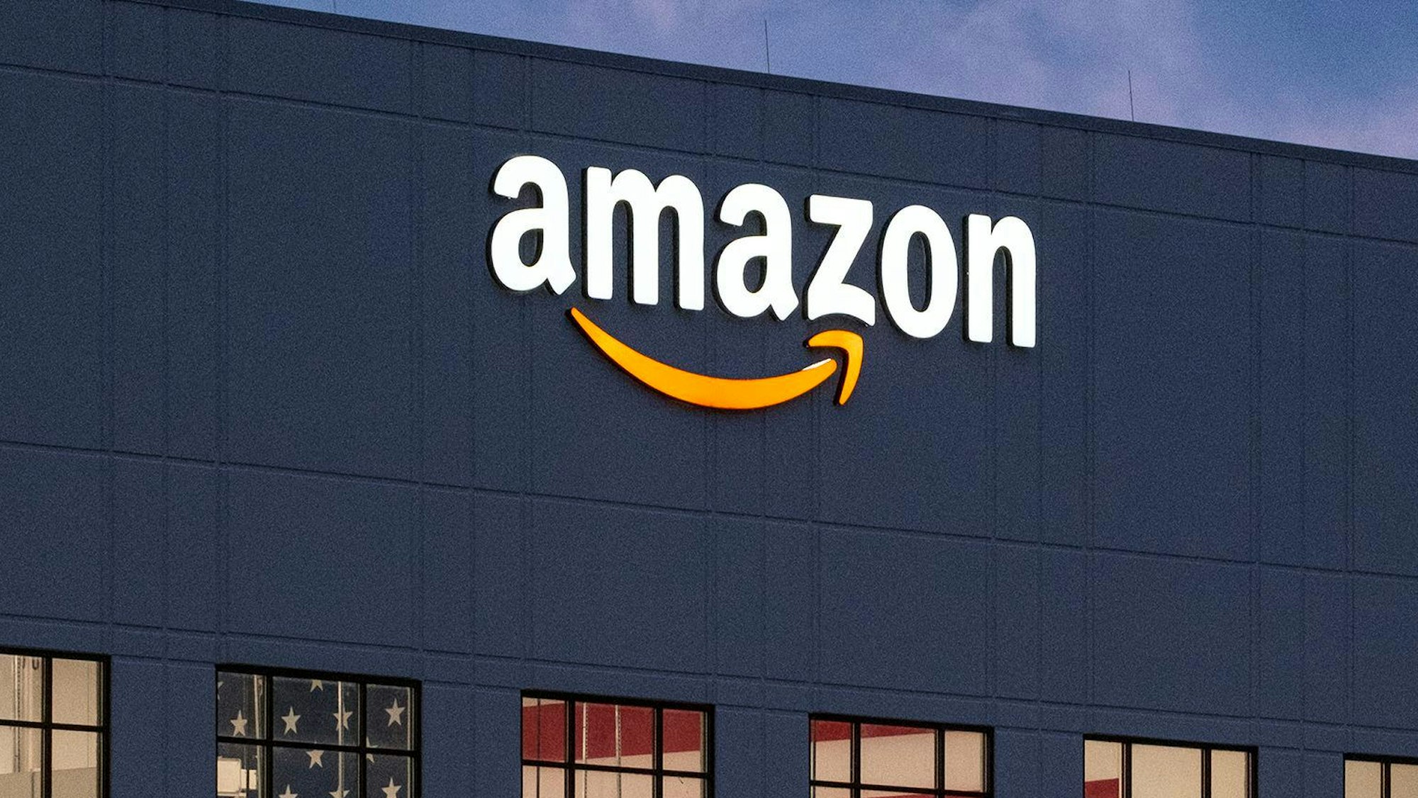 Zu sehen ist das Amazon-Logo auf einem Verteilzentrum des Unternehmens. Die Buchstaben leuchten weiß, darunter ist ein gelber Pfeil zu sehen. (Symbolbild)