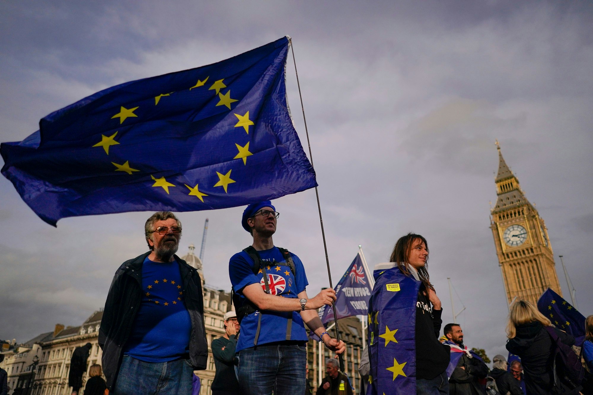 Vor dem britischen Parlament stehen mehrere Personen in blauen T-Shirts mit gelben Sternen und schwenken eine Fahne mit dem EU-Emblem.