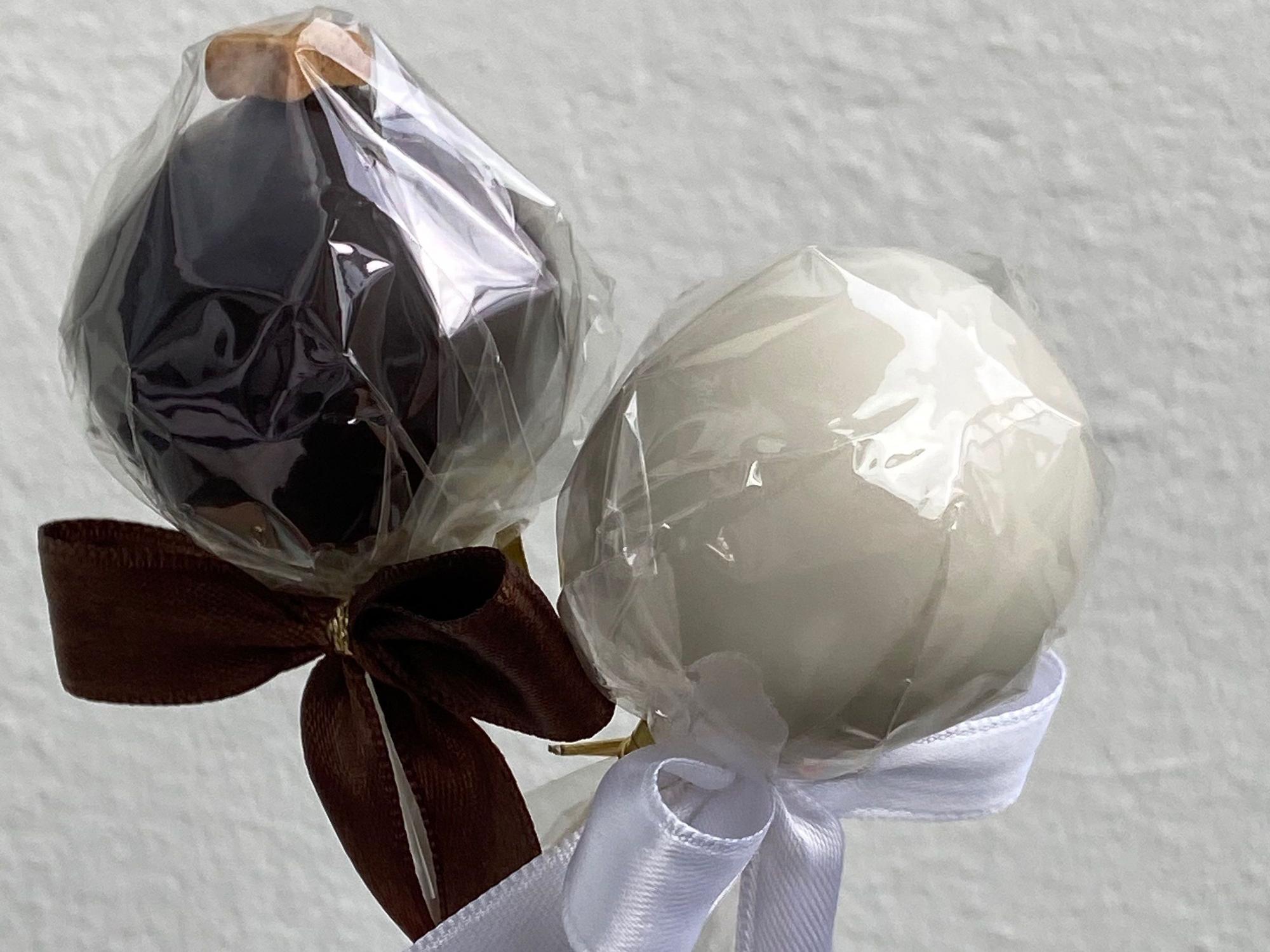 Zwei Cakepops sind in kompostierbarer Folie eingepackt und mit Schleifen zugebunden. Der linke Cakepop ist dunkelbraun, der rechte ist cremefarbend.