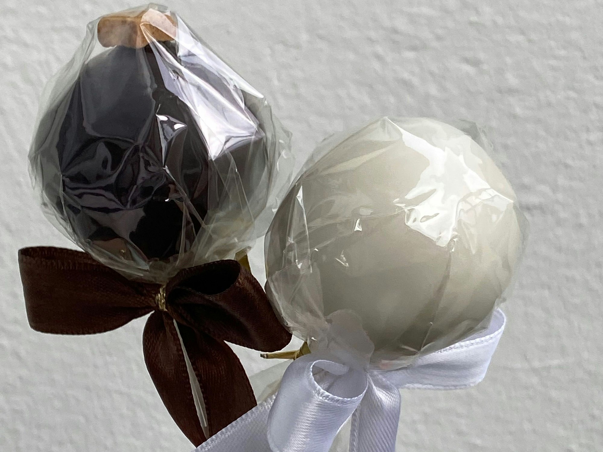 Zwei Cakepops sind in kompostierbarer Folie eingepackt und mit Schleifen zugebunden. Der linke Cakepop ist dunkelbraun, der rechte ist cremefarbend.