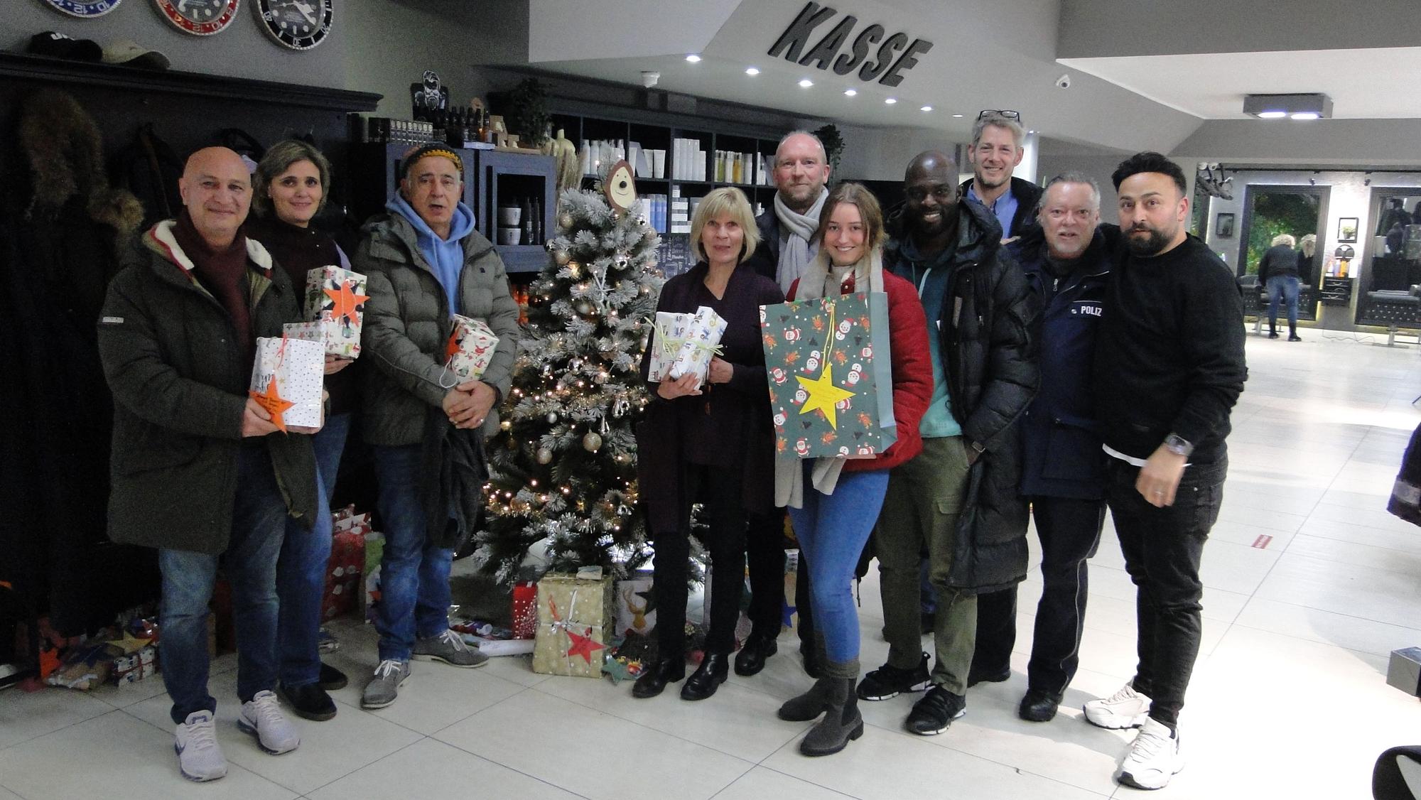 Zehn Personen stehen teils mit hübsch verpackten Geschenken in der Hand um einen Weihnachtsbaum herum, der in einem Friseur-Salon steht.