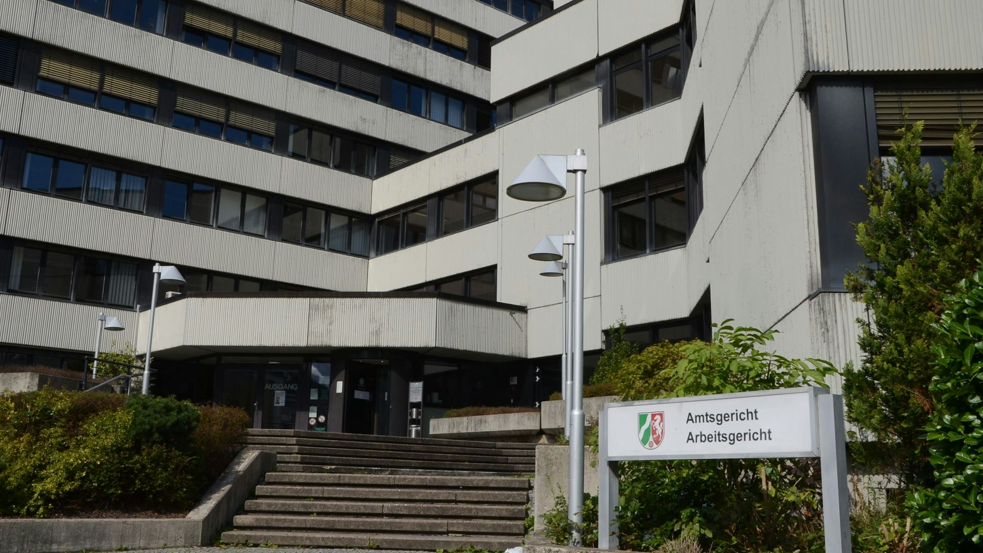 Im Hintergrund ist das Amtsgericht in Siegburg zu sehen. Vor dem Gebäude steht eine Hecke, daneben ein Schild, auf dem „Amtsgericht, Arbeitsgericht“ steht.