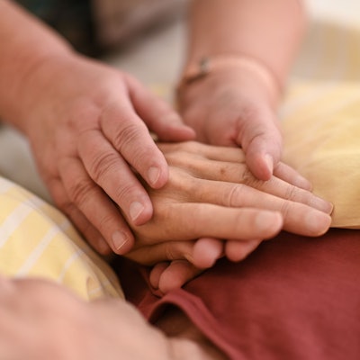 Eine Person hält mit beiden Händen die Hand eines Menschen, der mit einer gelben Decke zugedeckt ist.