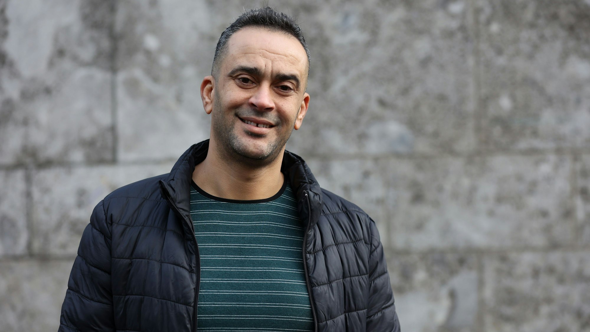 Ein Mann steht vor der Wand in einer blauen Übergangjacke und einem gestreiften Pillover.