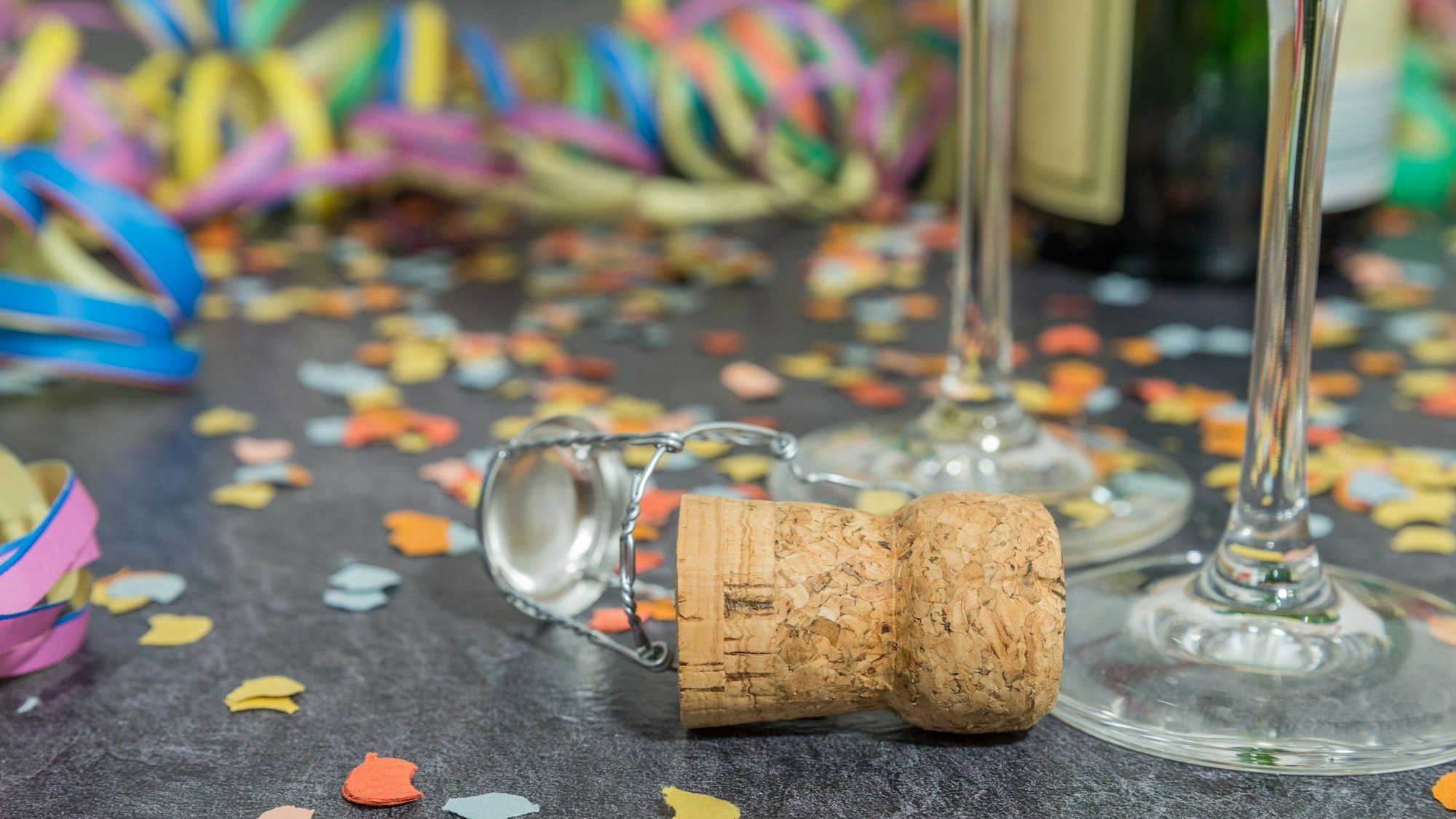 Ein Sektkorken liegt zwischen Konfetti, Luftschlangen und zwei Gläsern auf dem Tisch.