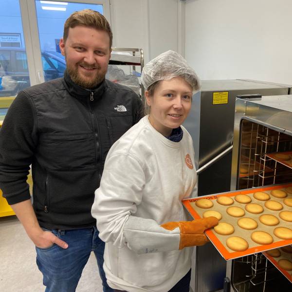 Kerstin Hottewitzsch und Nicolas Gritto stehen vor einem Industrie-Ofen. Hottewitzsch hält ein Blech mit Keksen und schiebt es in den Ofen. Sie lächeln beide in die Kamera.