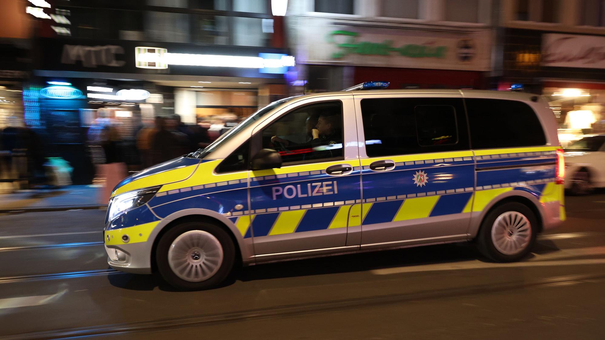 Ein Wagen der Polizei fährt über eine Straße, im Hintergrund sind Gebäude und Menschen verschwommen zu sehen.