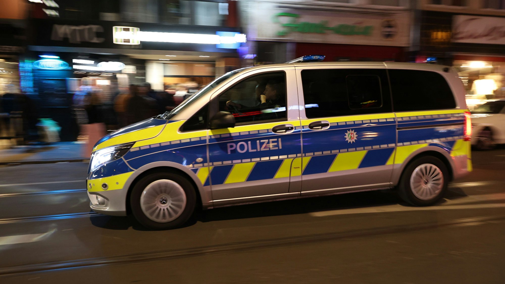 Ein Transporter der Polizei fährt nachts auf einer gut beleuchteten Straße durch eine Stadt. Der Hintergrund ist unscharf fotografiert.