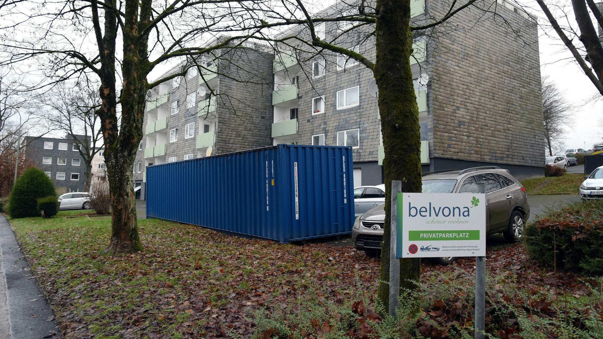 Blick auf ein Mehrfamilienhaus, davor ein Parkplatz und ein großer Müllcontainer.