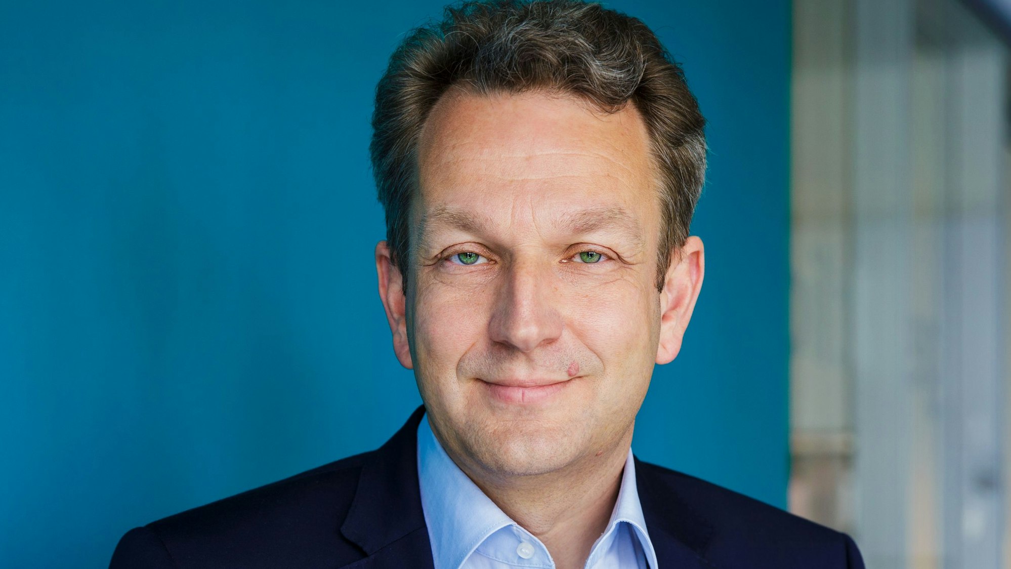 Ein Mann mittleren Alters mit blauem Sakko steht vor einer blauen Wand. Es ist Jan Schneck.