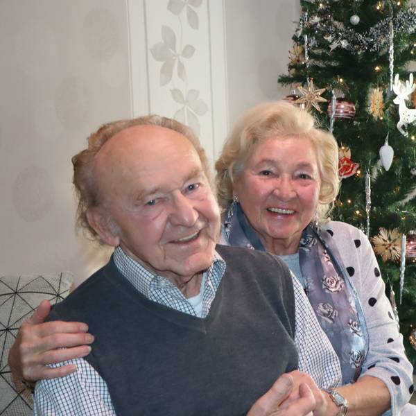 Ein älterer Mann und eine Frau sitzten vor dem Tannenbaum. Die Frau legt ihren Arm um seine Schulter.