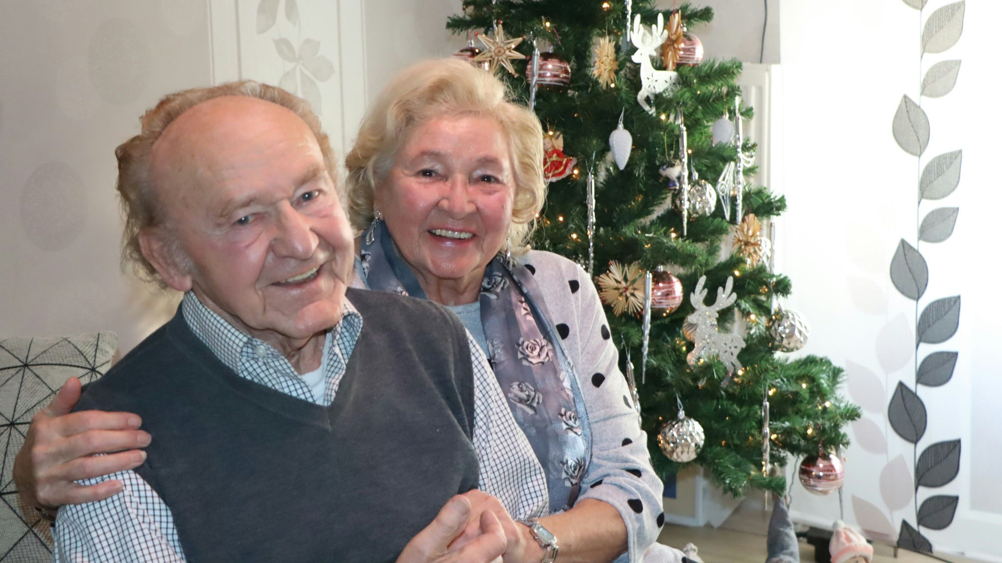 Ein älterer Mann und eine Frau sitzten vor dem Tannenbaum. Die Frau legt ihren Arm um seine Schulter.