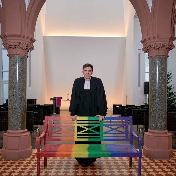 Tim Lahr, evangelischer Pfarrer, steht im Talar in der Kölner Christuskirche am Stadtgarten. Er stützt sich mit den Händen auf eine Holzbank, die in Regenbogenfarben gestrichen ist.