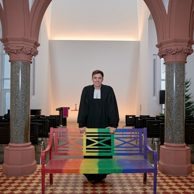 Tim Lahr, evangelischer Pfarrer, steht im Talar in der Kölner Christuskirche am Stadtgarten. Er stützt sich mit den Händen auf eine Holzbank, die in Regenbogenfarben gestrichen ist.