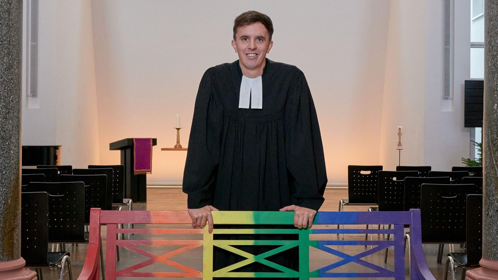 Tim Lahr, evangelischer Pfarrer, steht im Talar in der Kölner Christuskirche am Stadtgarten. Er stützt sich mit den Händen auf eine Holzbank, die in Regenbogenfarben gestrichen ist.