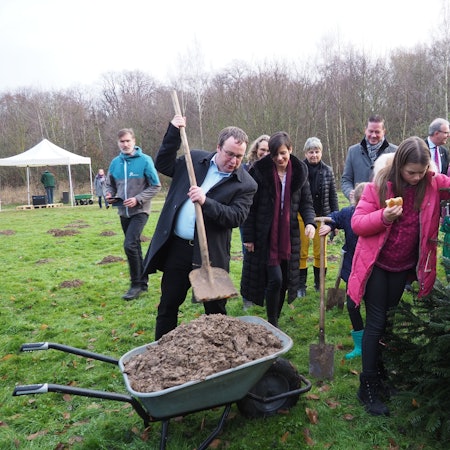 Viele Menschen stehen um einen Tannenbaum herum. Ein Mann schaufelt Erde aus einer Schubkarre.