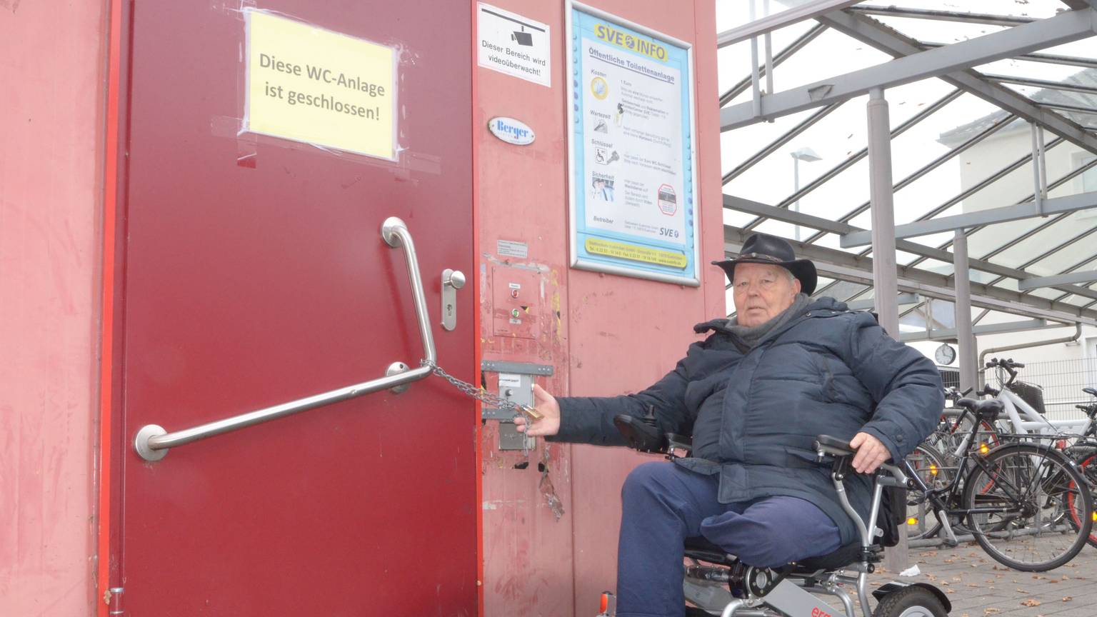 Der Rollstuhlfahrer Wilhelm Hachenberg vor einer geschlossenen Toilette am Bahnhof Euskirchen. An der Tür hängt eine Kette mit einem Vorhängeschloss.
