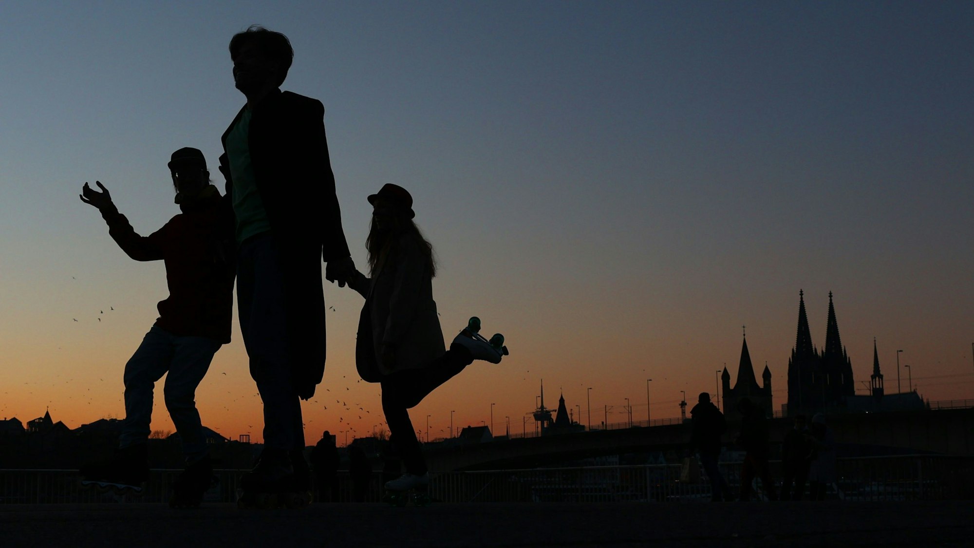 Im März tanzen drei Freunde auf Rollschuhen bei Dämmerung vor dem Stadtpanorama.