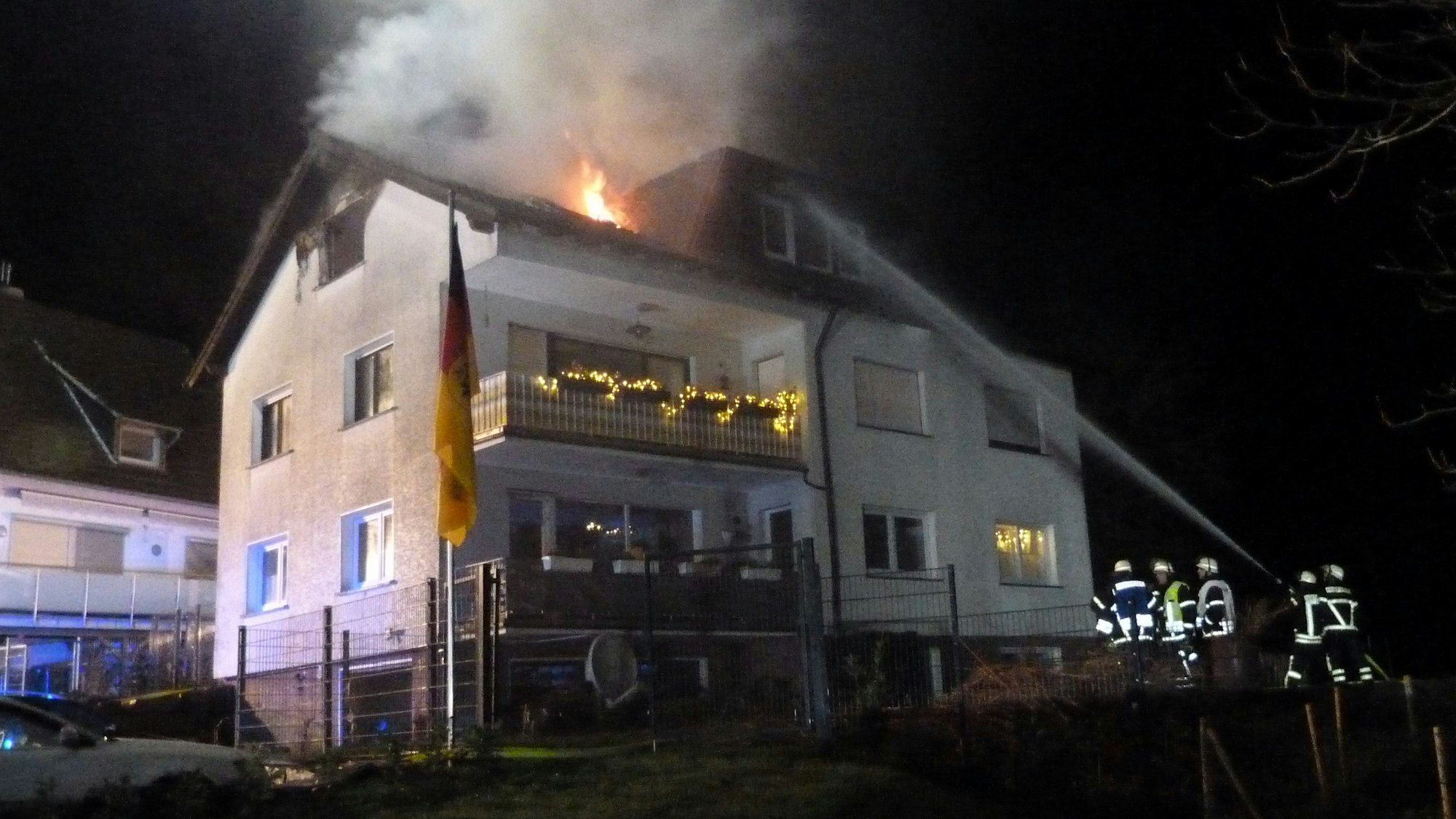 Aus dem Dach eines Hauses schlagen Flammen. Feuerwehrleute halten mit einem Löschwasser-Schlauch darauf.