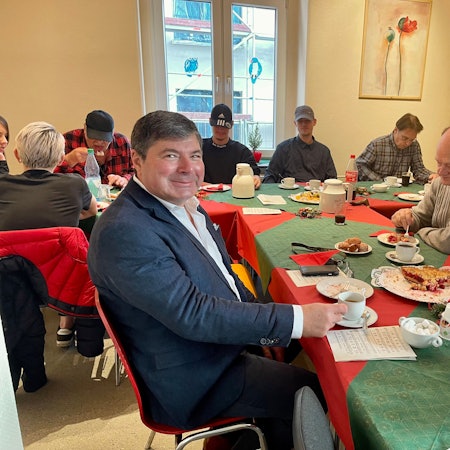 An einer langen Tafel sitzen mehrere Menschen vor Kaffeetassen und Kuchentellern.