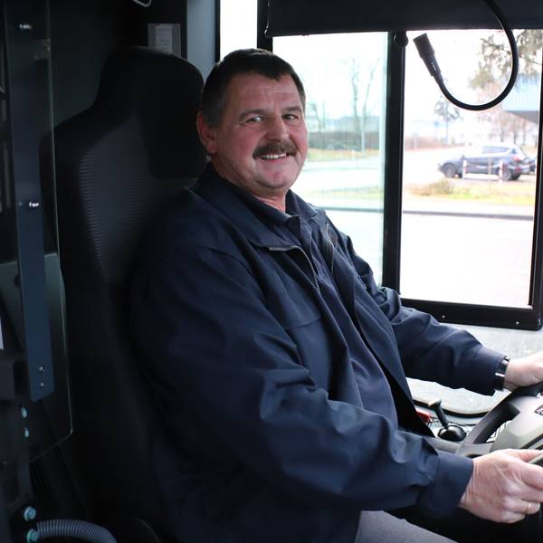 Ein Mann mit Schnurrbart sitzt am Steuer eines Busses, seine Hände halten das Lenkrad.