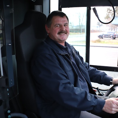 Ein Mann mit Schnurrbart sitzt am Steuer eines Busses, seine Hände halten das Lenkrad.