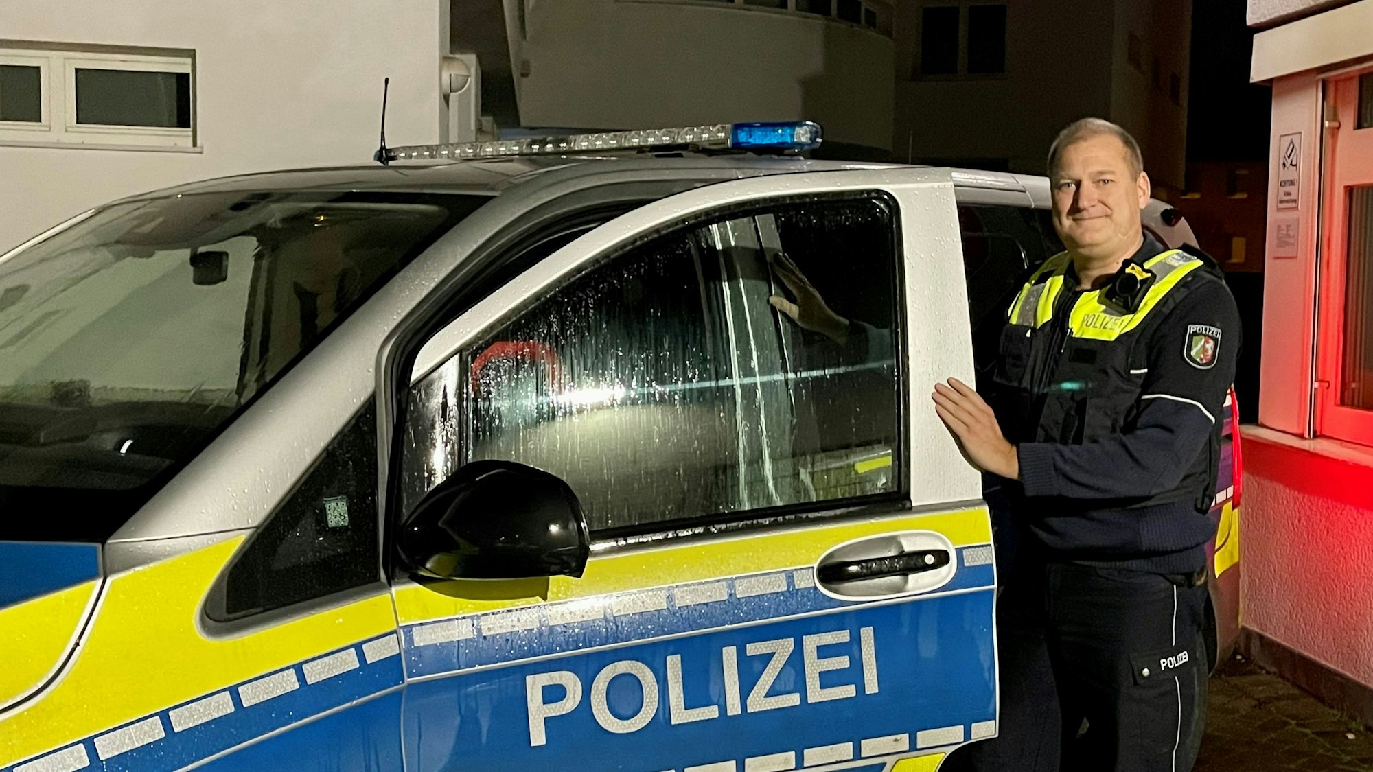 Ein Polizist in Uniform steht an der geöffneten Tür eines Streifenwagens. Es ist Nacht.