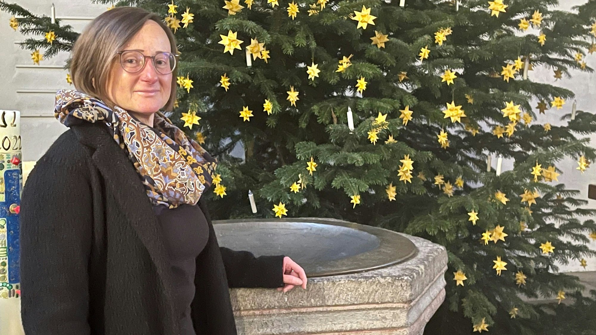Eine Frau mit Brille, großem Halstuch und Mantel steht an einem steinernen Taufbecken. Im Hintergrund steht ein geschmückter Christbaum.
