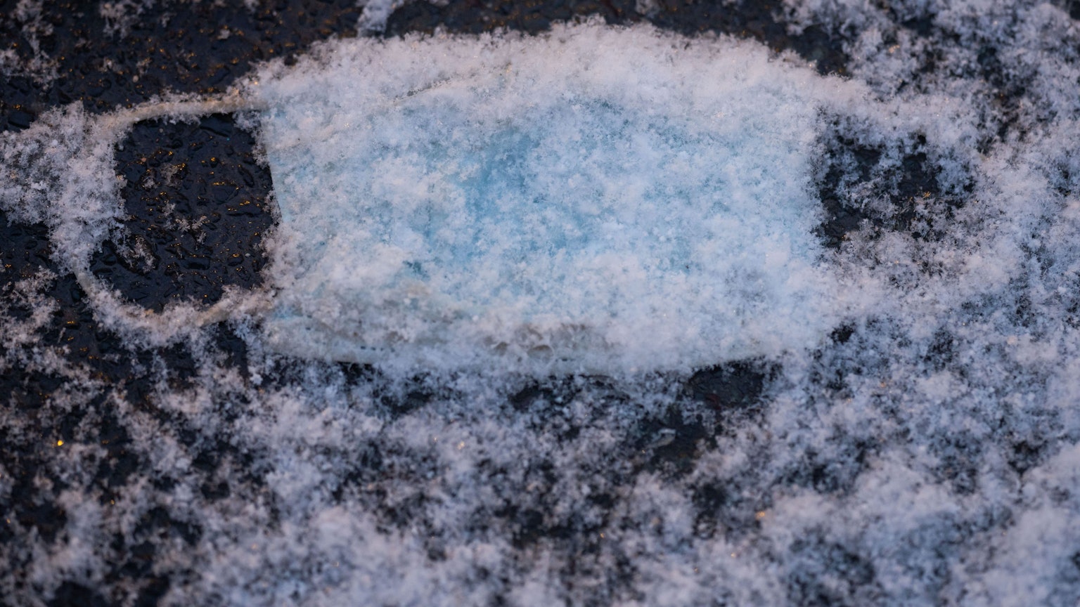Eine verlorene, medizinische OP-Maske als Mund-Nasenschutz liegt unter frisch gefallenem Schnee auf dem Pflaster.