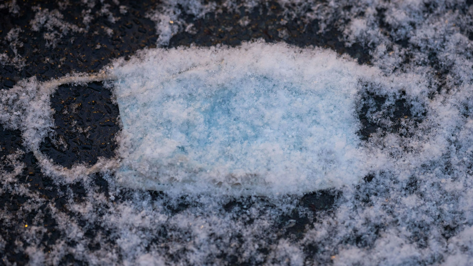Eine verlorene, medizinische OP-Maske als Mund-Nasenschutz liegt unter frisch gefallenem Schnee auf dem Pflaster.