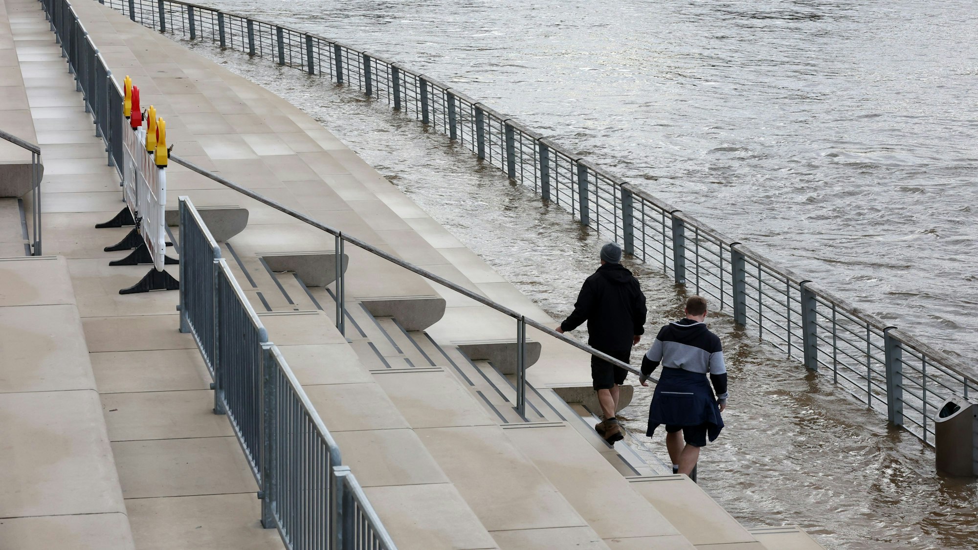Personen am Kölner Rheinboulevard während des kleinen Hochwassers, Warnbarken wurden bereits aufgestellt.
