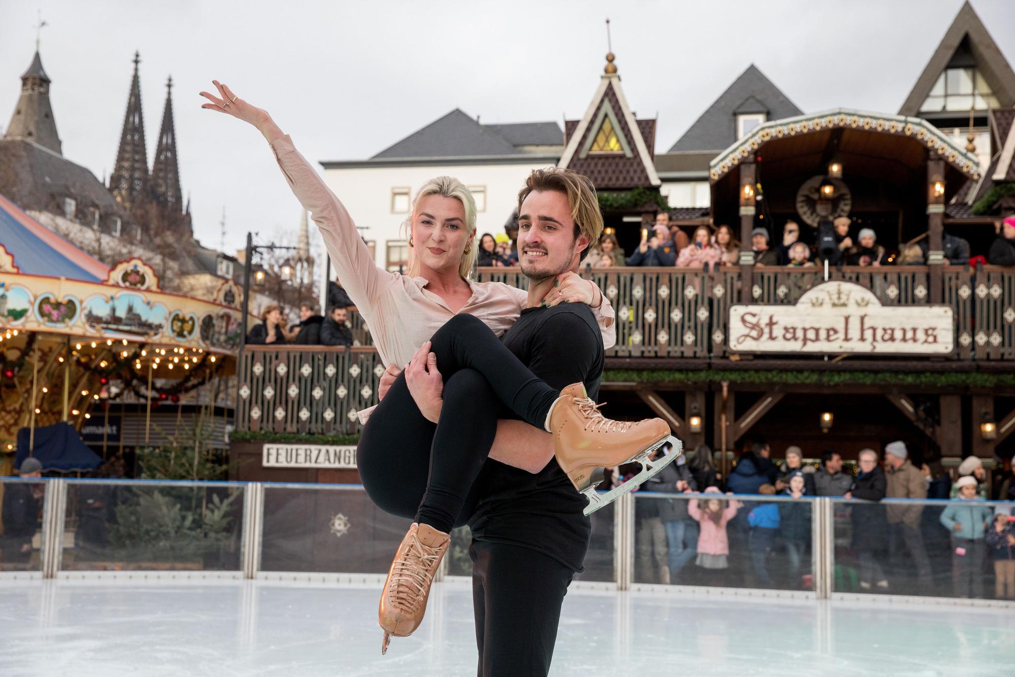 Tim Dieck hält Olivia Smart auf der Eisfläche elegant in beiden Armen. Im Hintergrund ist der Weihnachtsmarkt zusehen.