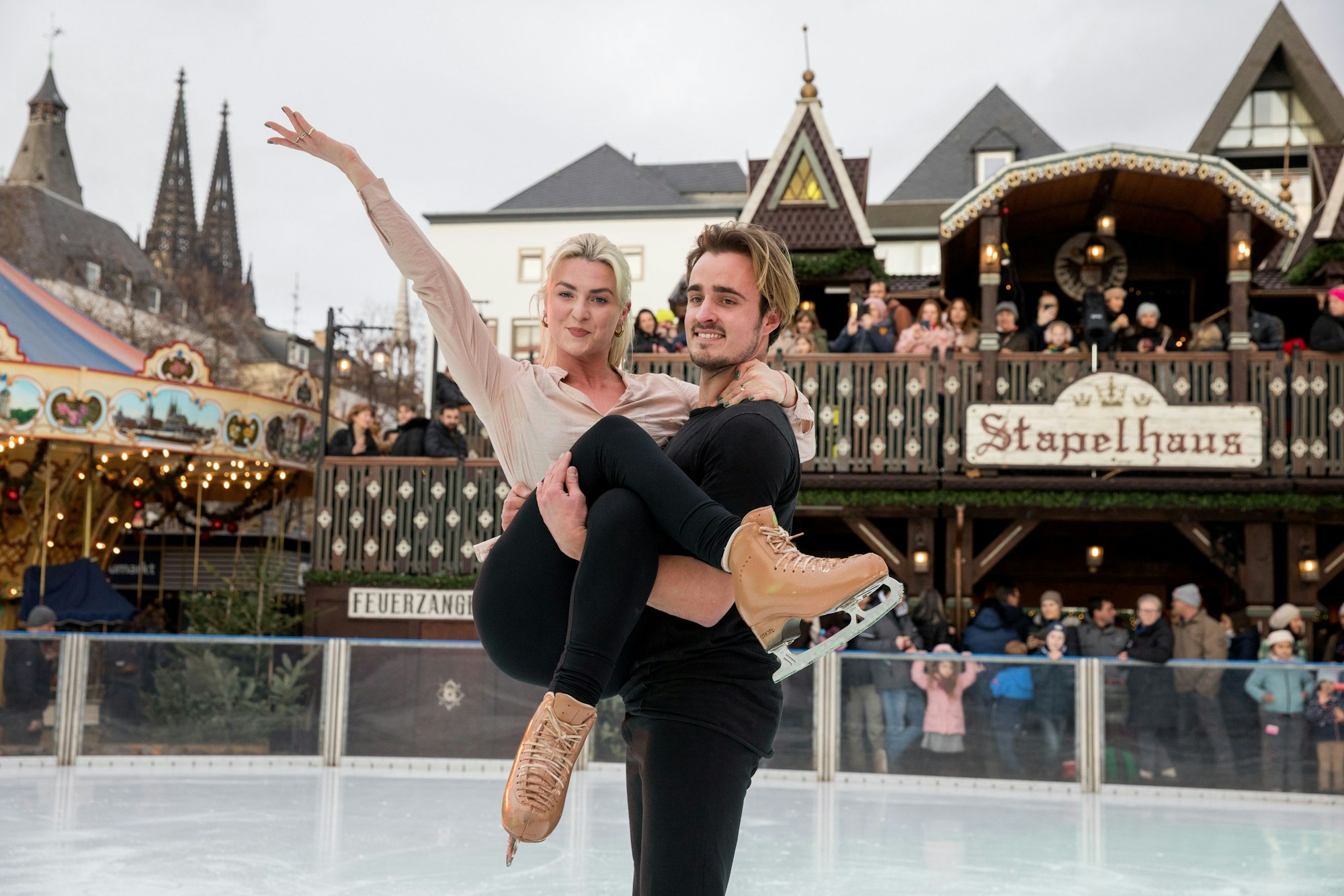 Tim Dieck hält Olivia Smart auf der Eisfläche elegant in beiden Armen. Im Hintergrund ist der Weihnachtsmarkt zusehen.
