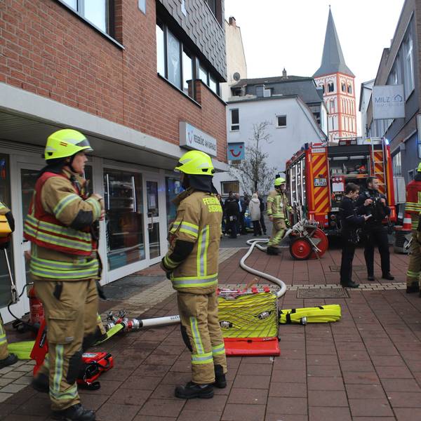Feuerwehrleute stehen auf dem gepflasterten Nogener Platz neben dem gläsernen Eingang. Ein dicker Schlauch liegt auf dem Boden bis zum Feuerwehrauto im Hintergrund.