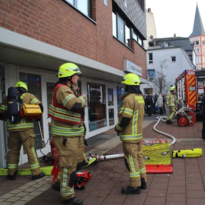 Feuerwehrleute stehen auf dem gepflasterten Nogener Platz neben dem gläsernen Eingang. Ein dicker Schlauch liegt auf dem Boden bis zum Feuerwehrauto im Hintergrund.